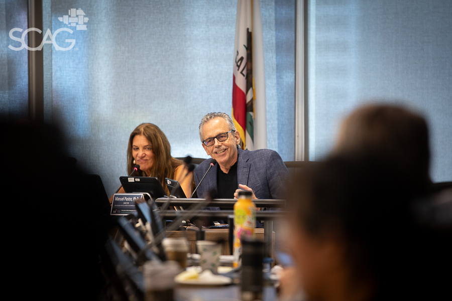 Man and woman at a conference table, smiling, with laptops and a flag behind.