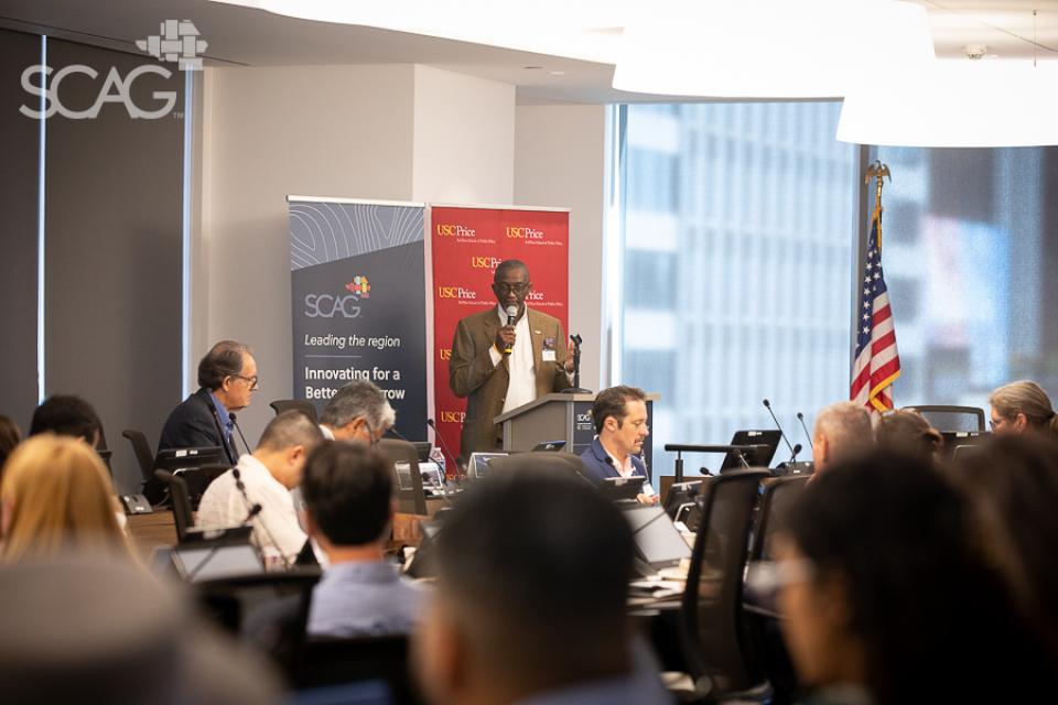 Man speaking at conference with seated audience, flags, and banners.