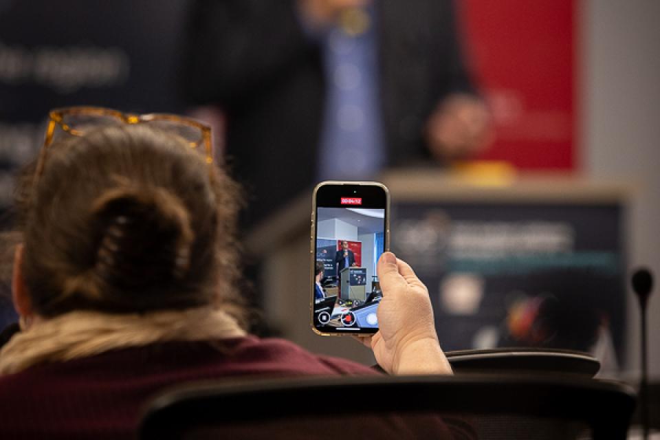 Audience member films speaker on phone in conference setting.